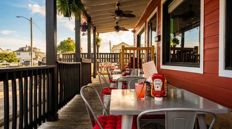 Outdoor restaurant patio with red siding, wooden deck, metal tables, and red cushioned chairs.