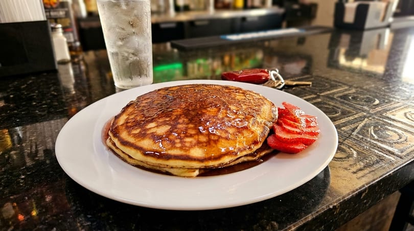 Stack of pancakes with syrup and sliced strawberries on a white plate at a bar.
